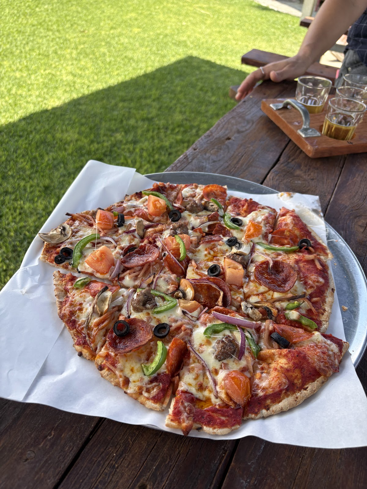 A loaded pizza on a timber table outside, with a tasting paddle behind it